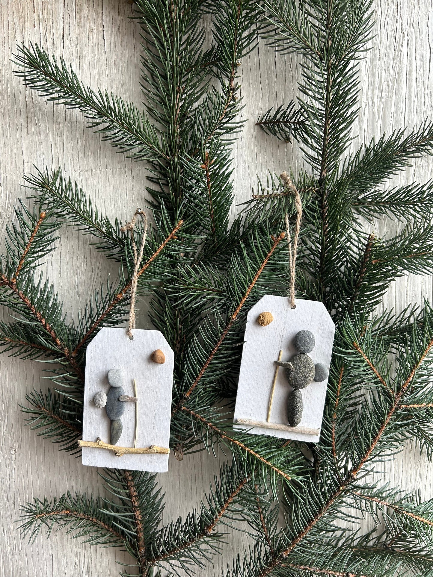 May include: Two rustic Christmas ornaments featuring hikers crafted from gray and white pebbles on small white wooden tags with twine hangers.  Each ornament depicts a simple hiker figure with a stick and a small round pebble, suggesting a sun or moon. The ornaments are displayed against a backdrop of dark green evergreen branches.