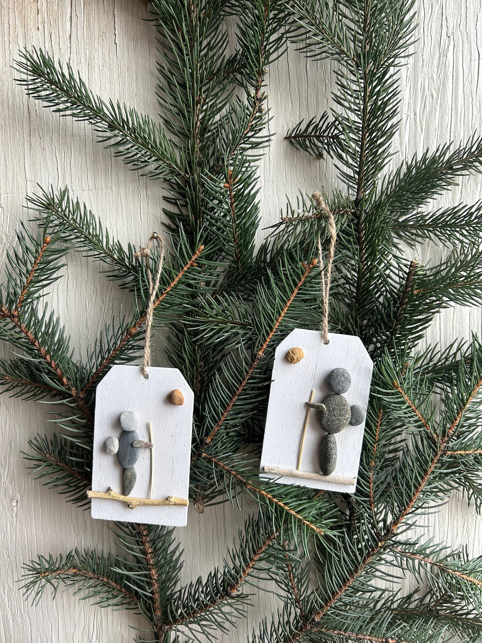 May include: Two rustic Christmas ornaments featuring hikers crafted from gray and white pebbles on small white wooden tags with twine hangers.  Each ornament depicts a simple hiker figure with a stick and a small round pebble, suggesting a sun or moon. The ornaments are displayed against a backdrop of dark green evergreen branches.