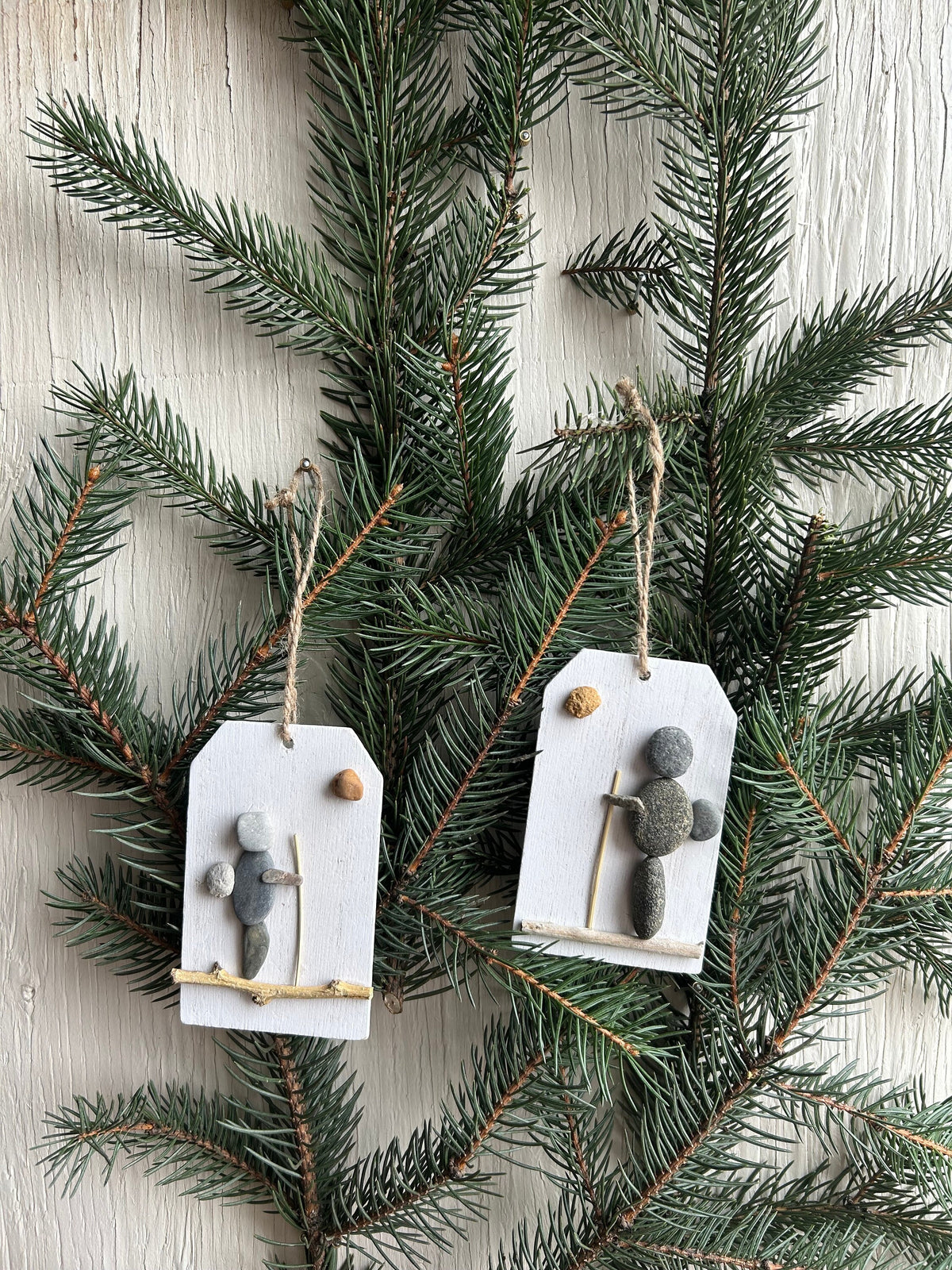 May include: Two rustic Christmas ornaments featuring hikers crafted from gray and white pebbles on small white wooden tags with twine hangers.  Each ornament depicts a simple hiker figure with a stick and a small round pebble, suggesting a sun or moon. The ornaments are displayed against a backdrop of dark green evergreen branches.