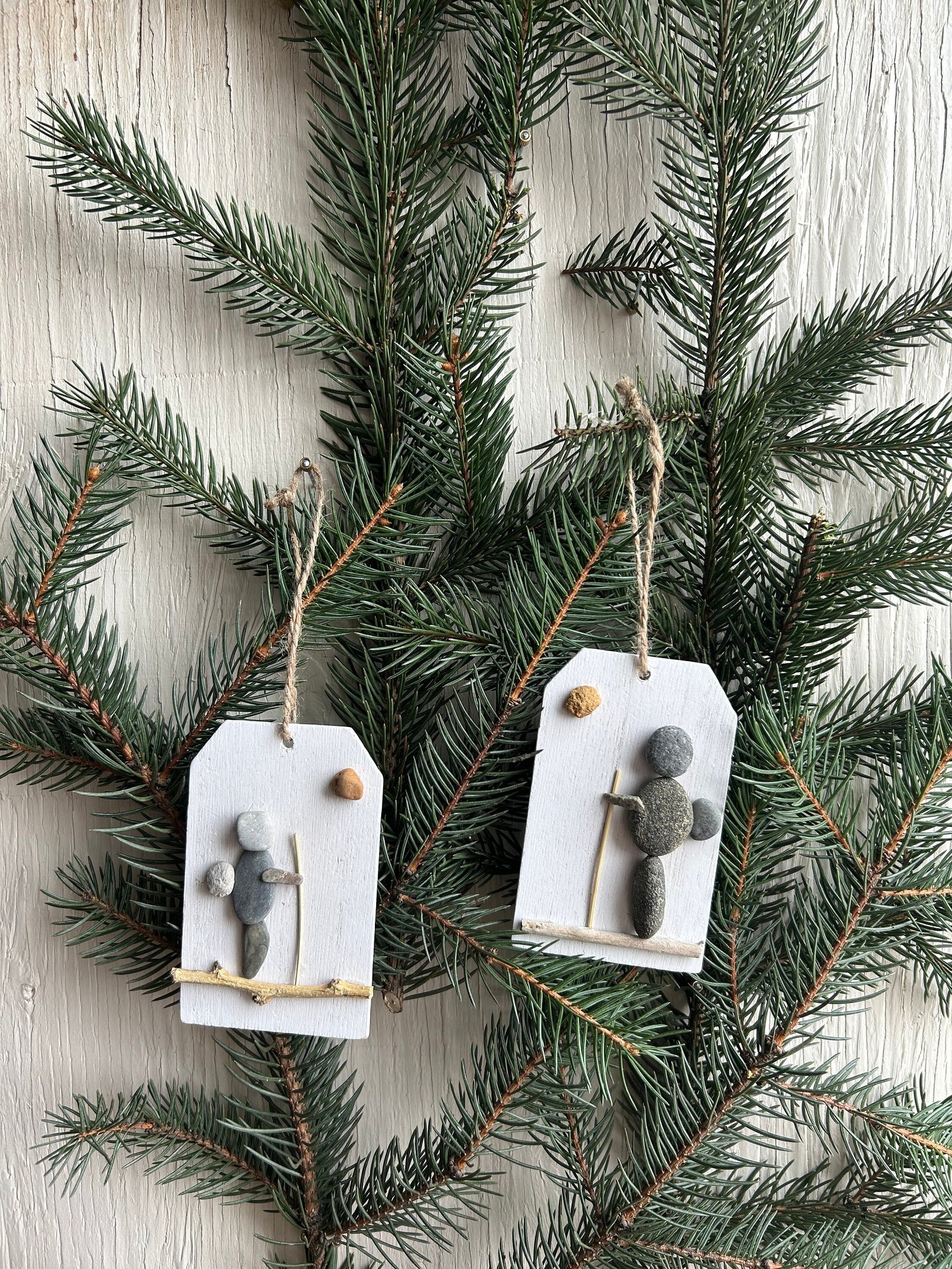 May include: Two rustic Christmas ornaments featuring hikers crafted from gray and white pebbles on small white wooden tags with twine hangers.  Each ornament depicts a simple hiker figure with a stick and a small round pebble, suggesting a sun or moon. The ornaments are displayed against a backdrop of dark green evergreen branches.