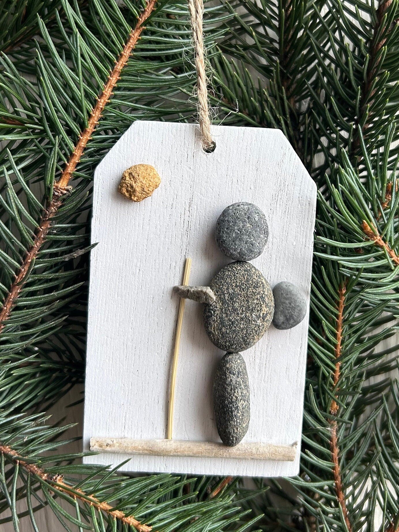 May include: Stone pebble Christmas ornament.  A gray, pebble person figurine stands on a small, light-colored wooden base. The figurine holds a small, light-colored stick. A small, tan-colored stone is positioned above the figurine. The ornament is attached to a piece of twine. The ornament hangs from a fir tree branch.