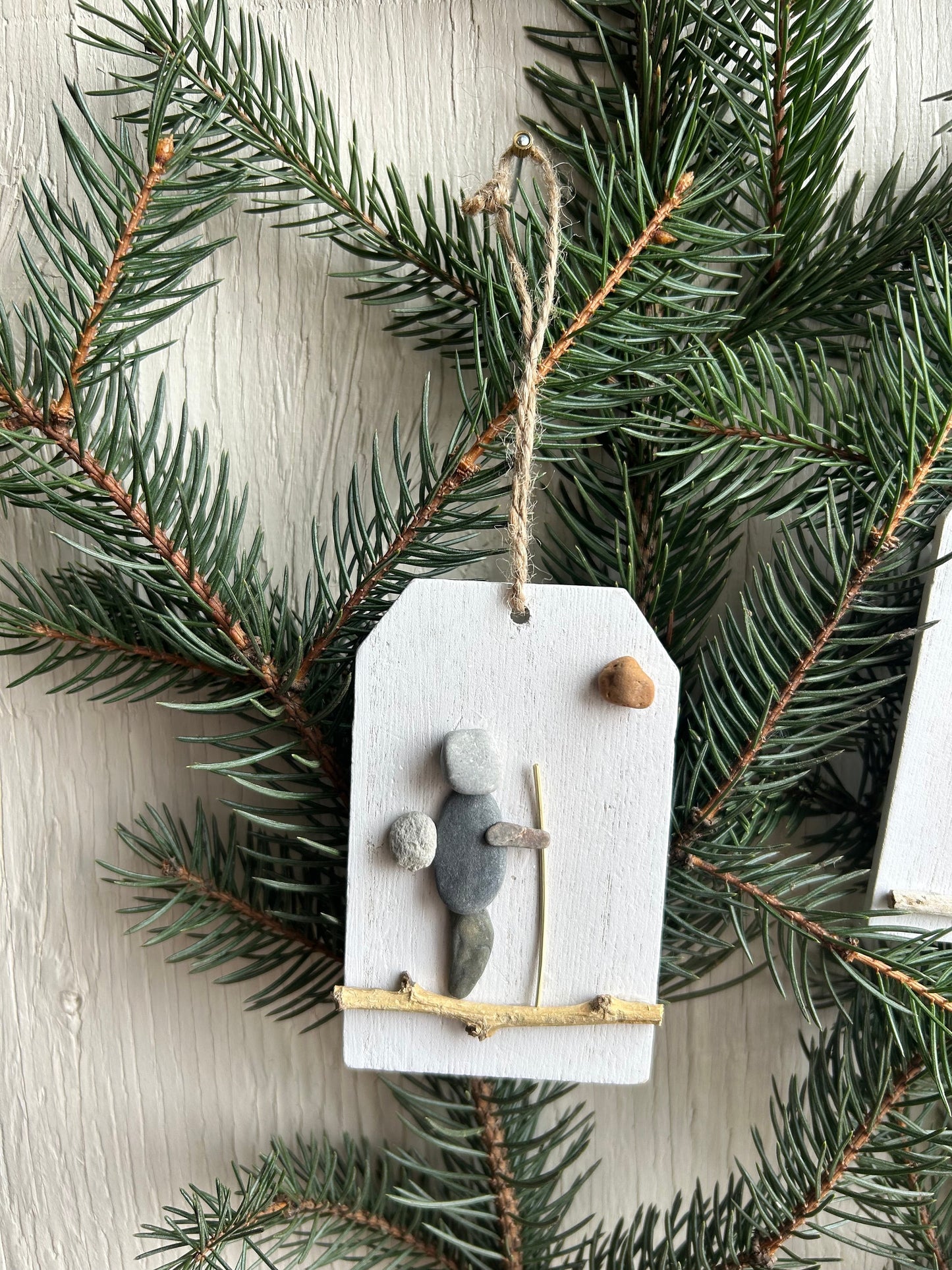 May include: Stone pebble art Christmas ornament.  A hiker figure is created using gray and white stones on a small white wooden tag. The hiker carries a small stone and a stick. A small brown stone is in the upper right corner. The ornament hangs from a twine string. The tag is accented with a small twig.  The ornament is displayed against a background of evergreen branches.