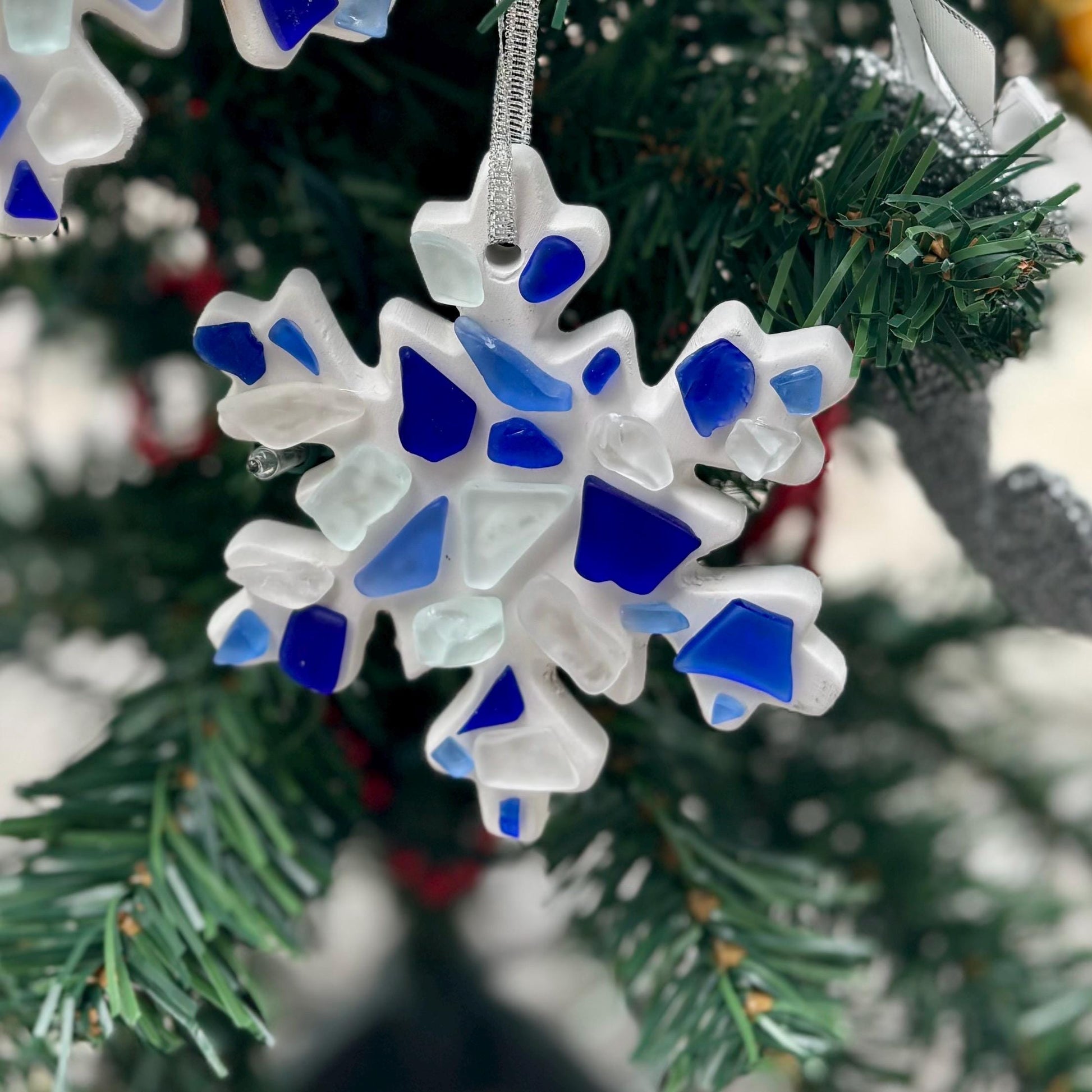 May include: White snowflake Christmas ornament decorated with blue and white sea glass pieces. The snowflake is made of resin or a similar material and has a silver cord for hanging.  The sea glass pieces are various shades of blue and white, creating a wintery design. This handmade ornament is perfect for adding a coastal touch to your holiday decor.