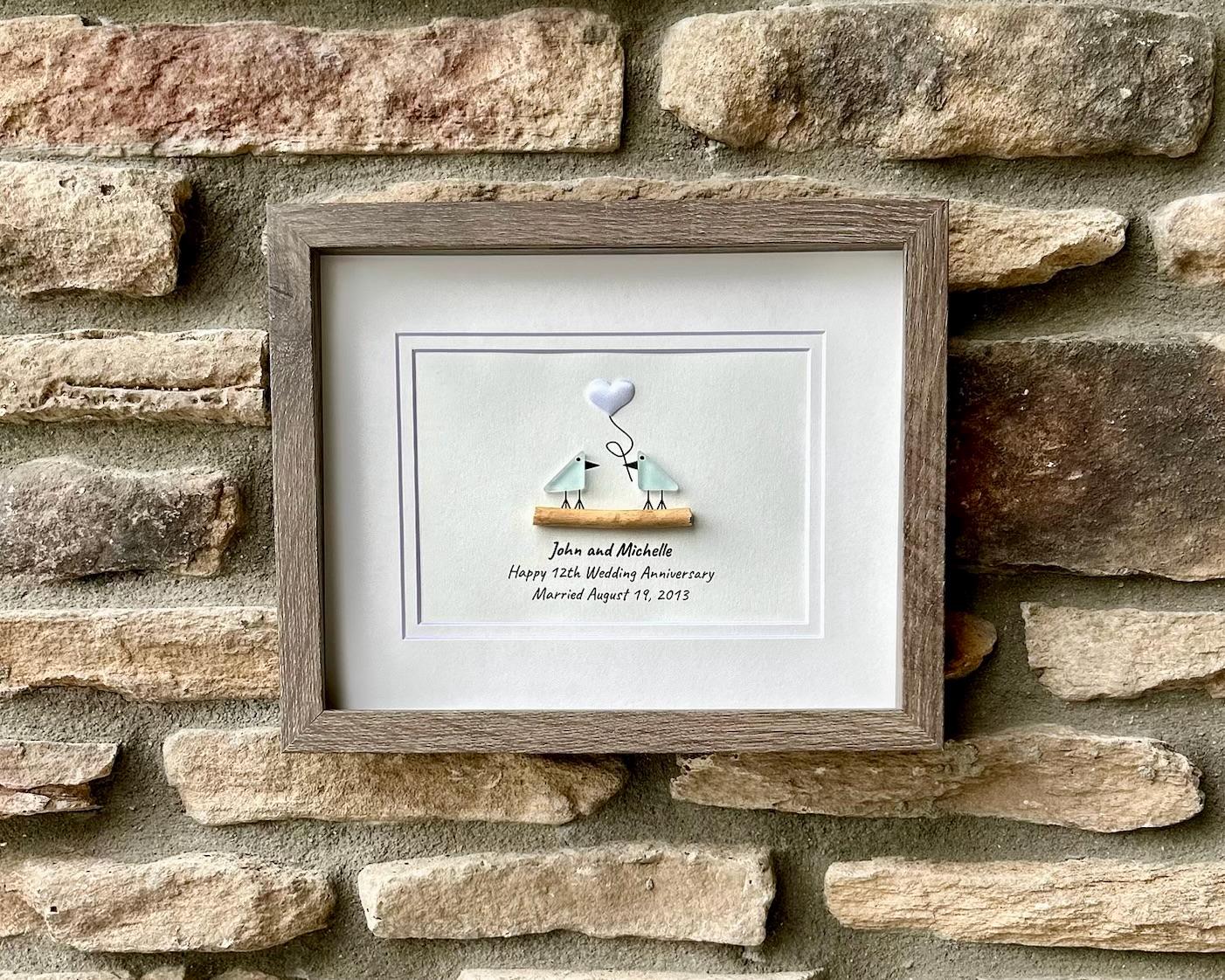 May include: A framed artwork featuring two light blue birds perched on a wooden branch. A white heart-shaped object is suspended above them. The artwork includes the text 'John and Michelle Happy 12th Wedding Anniversary Married August 19, 2011' in black font. The frame is a light brown color.