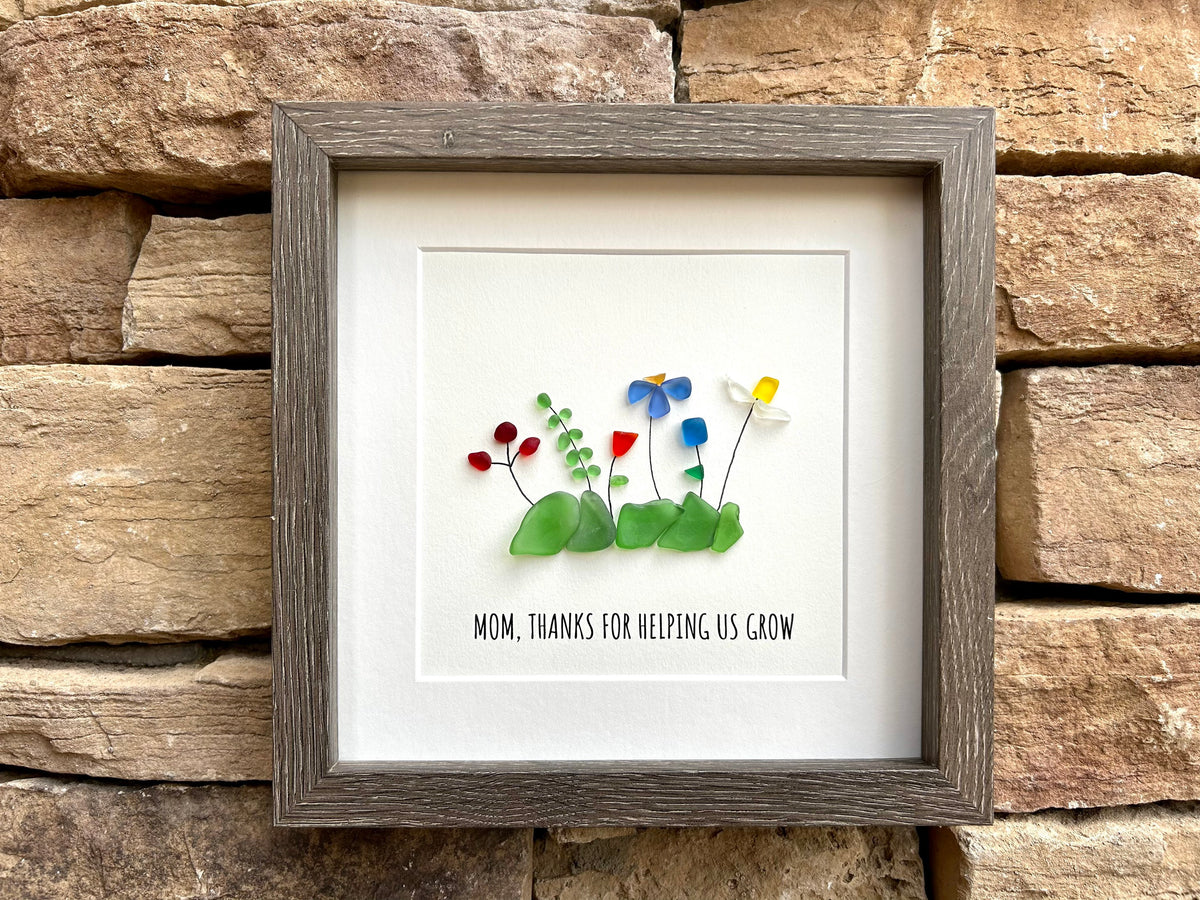 May include: A framed piece of art with a gray frame. The art is made of sea glass and features a variety of flowers in blue, red, yellow, and white. The text 'MOM, THANKS FOR HELPING US GROW' is written below the flowers.