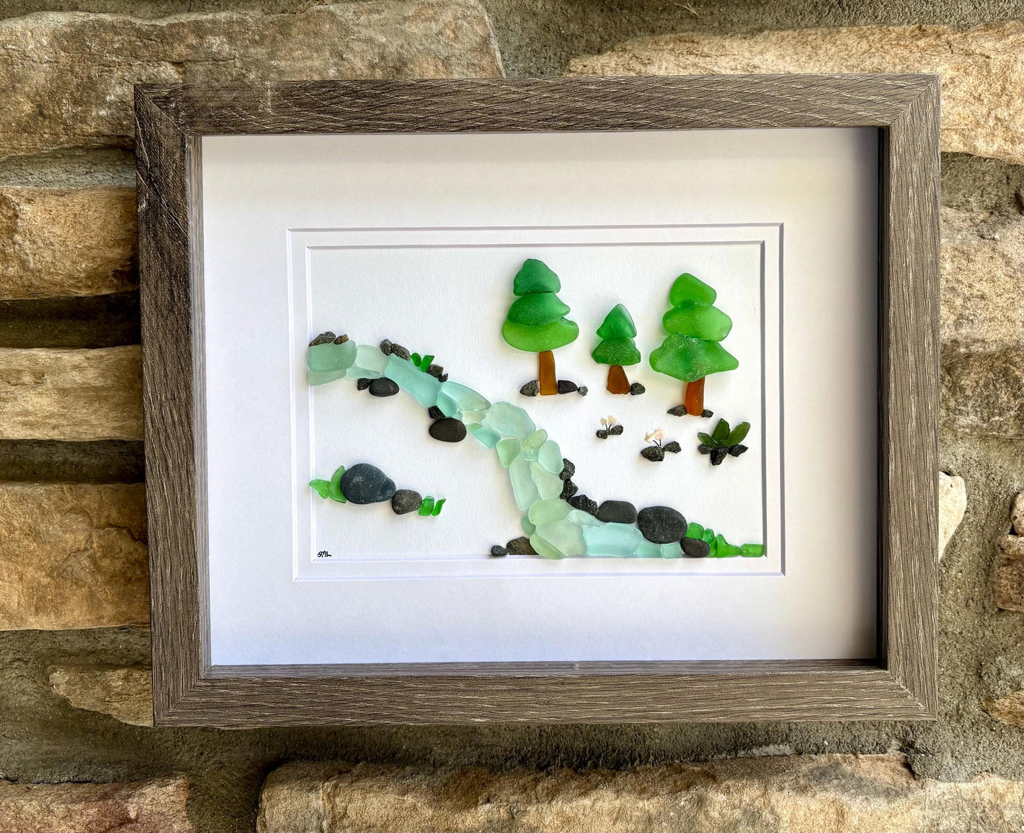 May include: Framed artwork depicting a serene landscape. A waterfall of blue and clear glass flows through the scene, with green glass trees and dark stones. The artwork is set in a gray wooden frame, mounted on a stone wall.